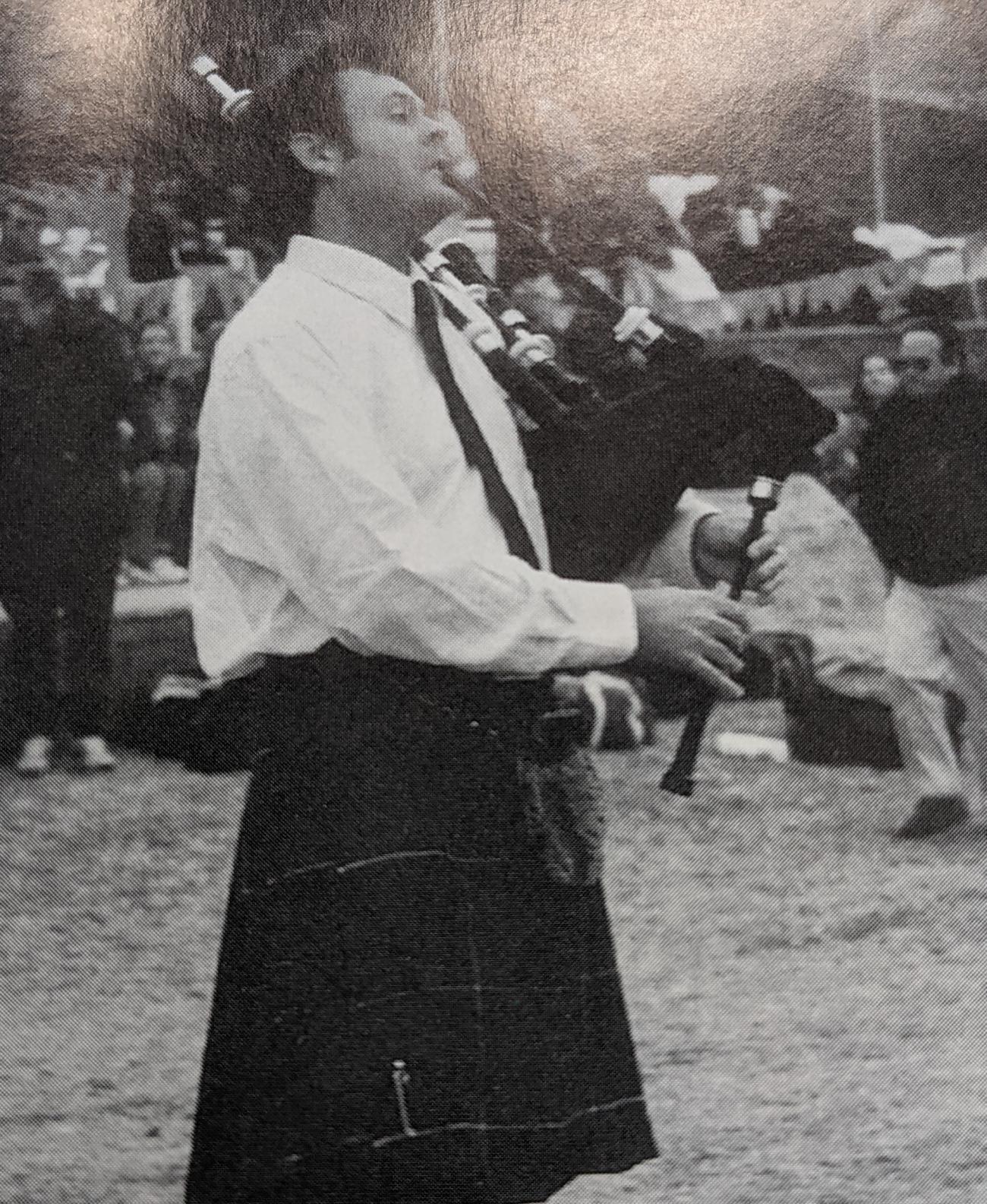 A black and white photo of piper wearing a kilt performing on a rugby pitch.